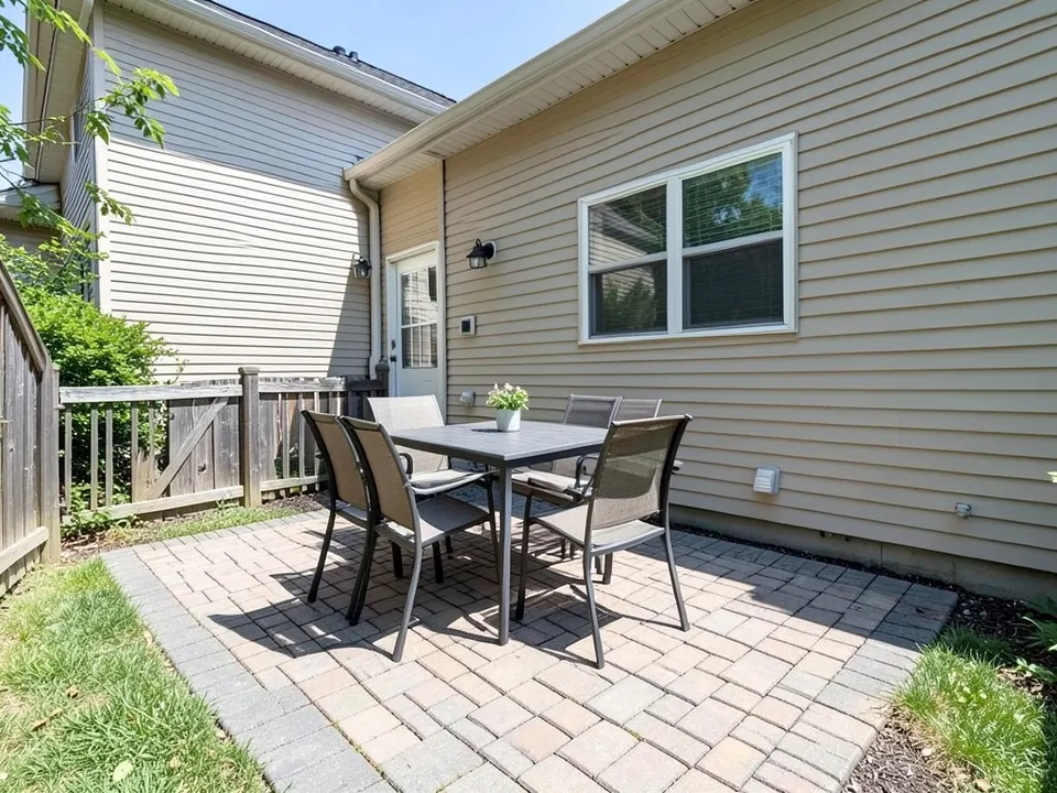 AI-generated example: a sunny paver patio with a table, four chairs and a flower pot, a beige house wall with a window and a door, a taller wall on the left with a wall lamp, a wooden fence and shrubs