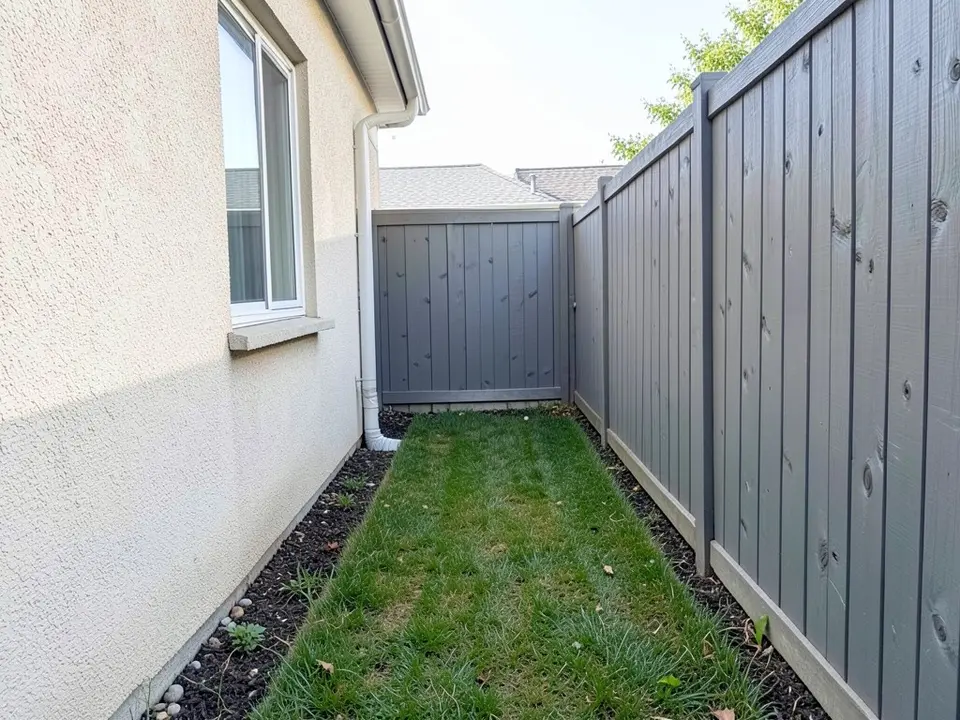 AI-generated example: a narrow side yard between a cream stucco house wall with a window and a white downspout on the left and a tall grey painted wooden fence on the right, a mown strip of grass with a dark mulch strip along the wall, and a grey fence and gate at the far end