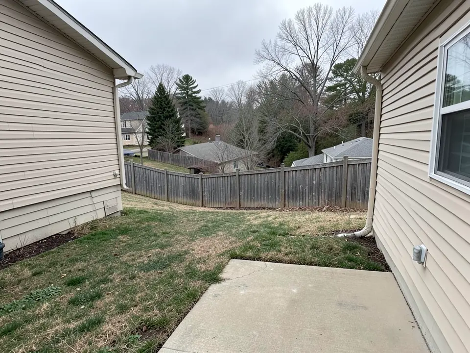 AI-generated example: a small concrete patio between two beige vinyl-sided houses, with a patchy lawn that slopes down to a weathered wooden privacy fence and neighbors' roofs and bare trees lower down beyond it