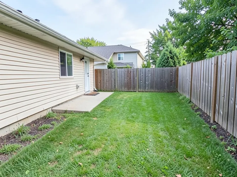 AI-generated example: a small backyard with a lawn, a beige-siding one-story house on the left with a small concrete slab at its door, a wooden fence at the back and along the right, and a dark mulch strip with a few plants beside the house and the fence