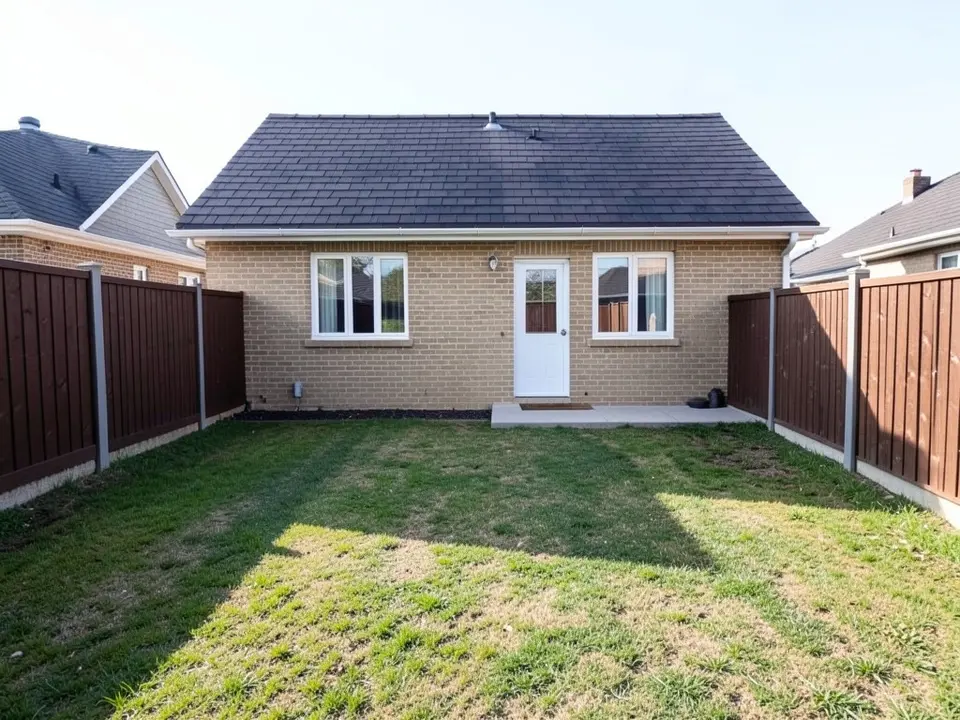 Narrow backyard behind a single-story tan brick house with a plain mowed lawn, brown wooden fences and a small concrete slab by the back door
