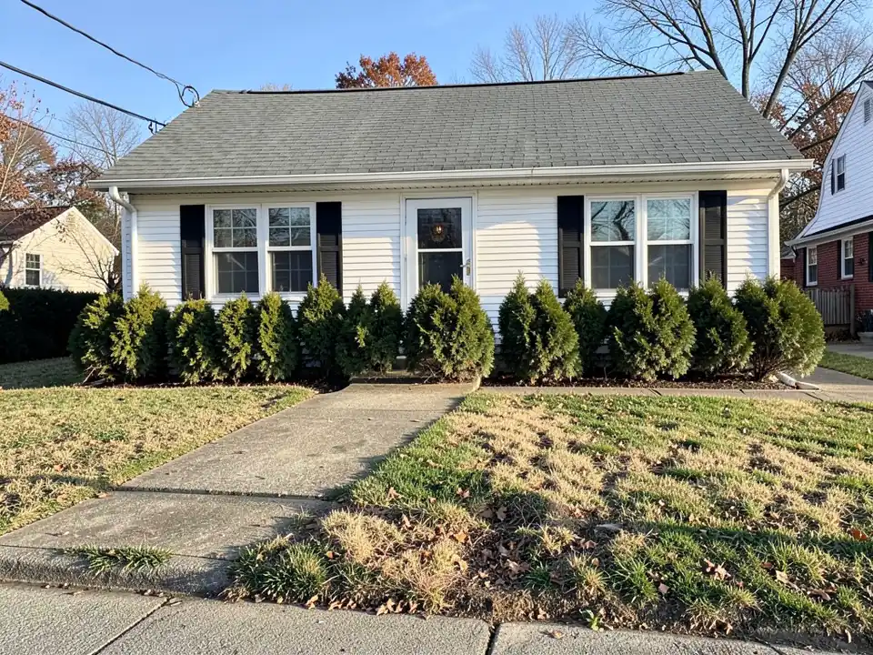 Small white house with black shutters, a row of overgrown evergreen shrubs hiding the front wall and a weedy, uneven lawn