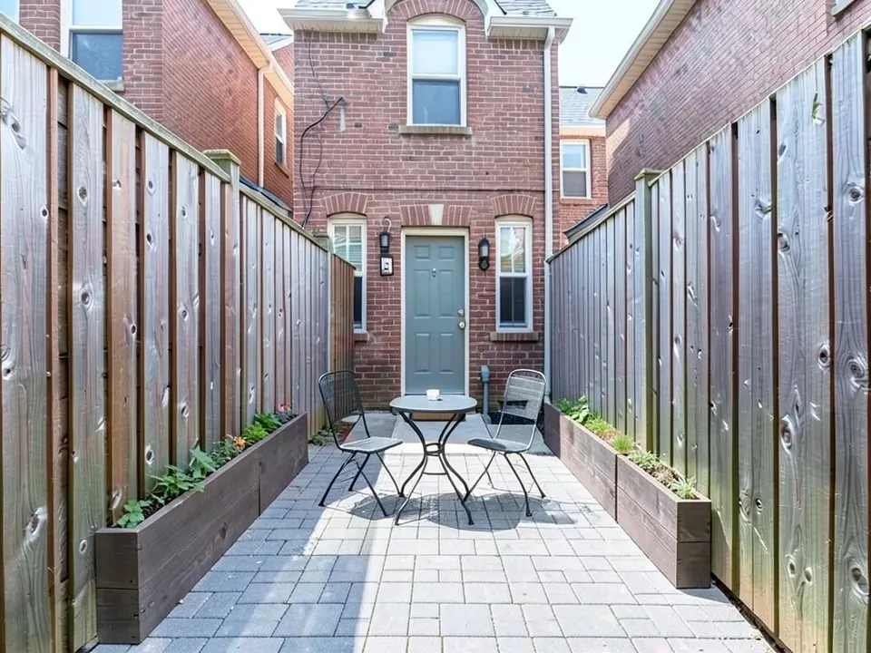 AI-edited version of the same image: the slab and lawn replaced by a grey paver patio with a small round metal bistro table and two metal chairs, and narrow wooden raised planters with green plants along both fences