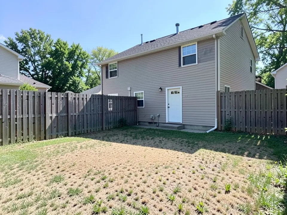 AI-generated example: the back of a two-storey beige house with a white back door and a wooden fence on both sides, over a patchy dry lawn with brown areas, in bright sun